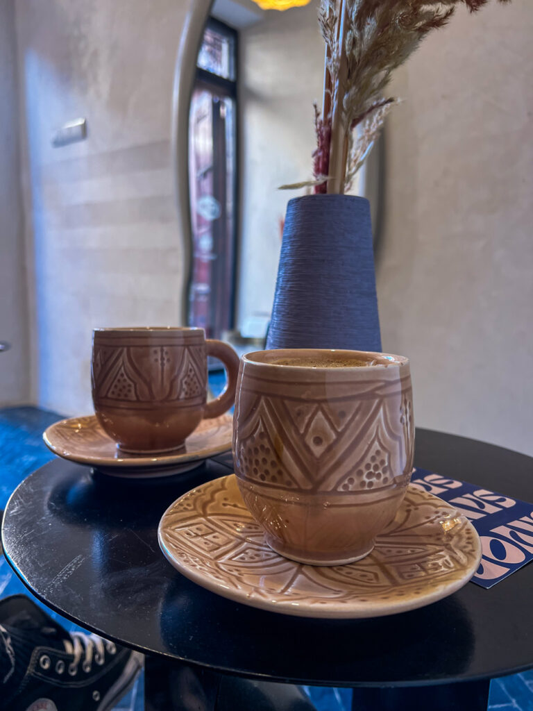 Two ceramic mugs with intricate Moroccan designs, filled with coffee, placed on a small black table with a decorative vase in the background.