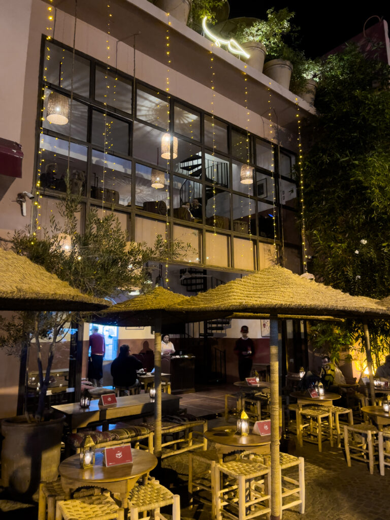 An outdoor dining area at night with straw canopies, warm lighting, and a modern building facade decorated with string lights at a Marrakesh restaurant.
